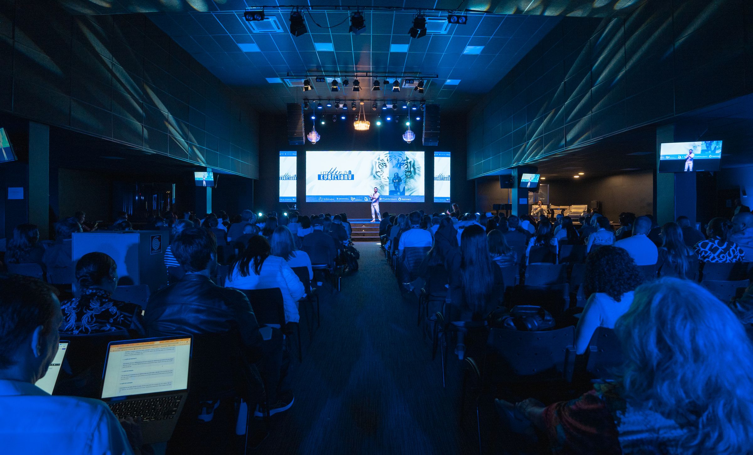 Auditório lotado visto do fundo da sala, com Du Oliva no palco ao fundo